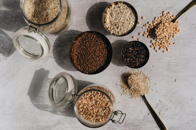 A top view of assorted grains and pulses in jars and bowls on a textured surface with shadow play.
